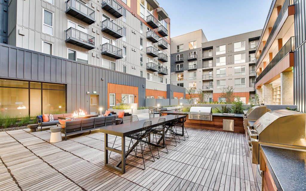 Nighttime view of the courtyard with path lighting, lounge seating and tables, a lit fire table, grilling stations and a high-top dining table with bar stools.
