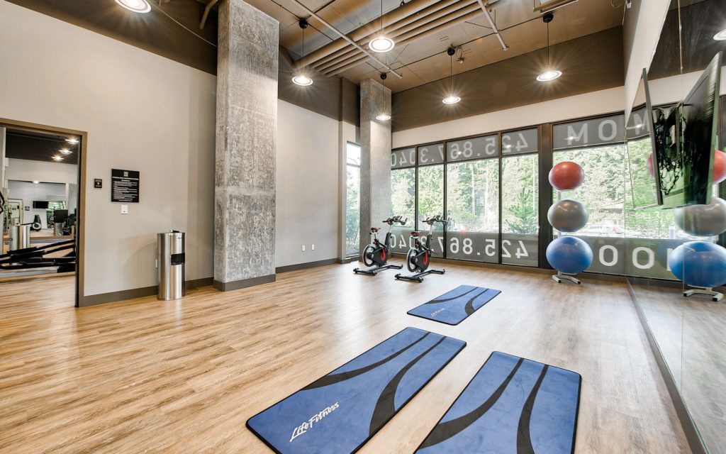 Fitness and yoga studio with wood flooring, mirrored wall with wall mounted TV, exposed pipe ceiling, stationary bikes, and yoga mats and balls.